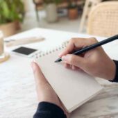 Closeup imagen de una mano escribiendo en un cuaderno en blanco con taza de café y postre en la mesa de café